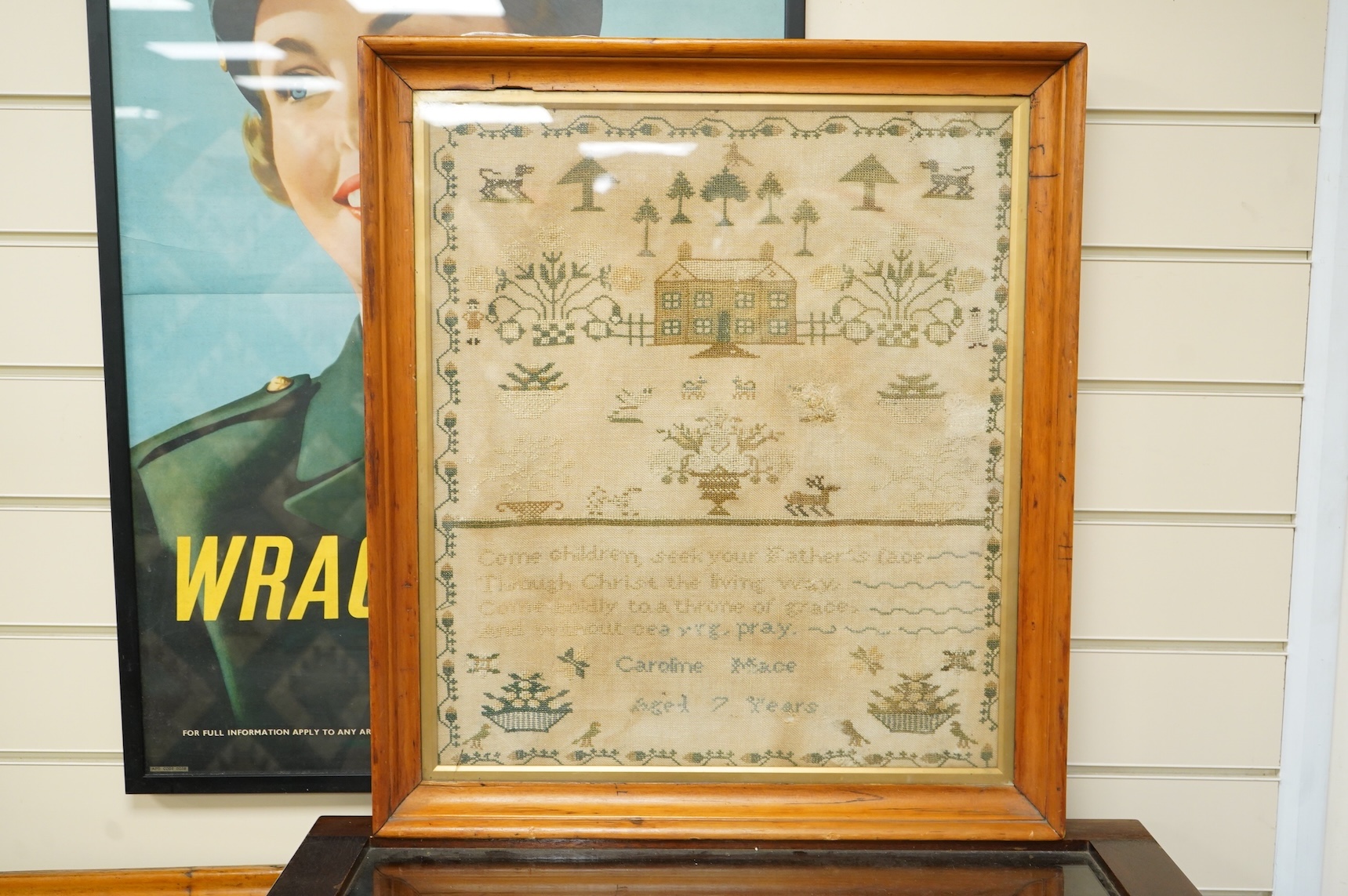 A framed 19th century sampler by Caroline Mace aged 7 years. Worked in cross stitch with the top half showing a scene of a house and trees, the lower half with a verse and various spot motifs within a vining border. 48.5